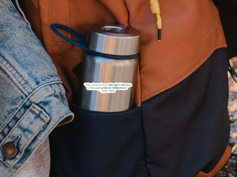 Image shows a person's backpacking gear and a water bottle with a Hand holding an John Muir naturalist sticker for the outdoor lovers. The quote reads, "The clearest way into the universe is through a forest wilderness."